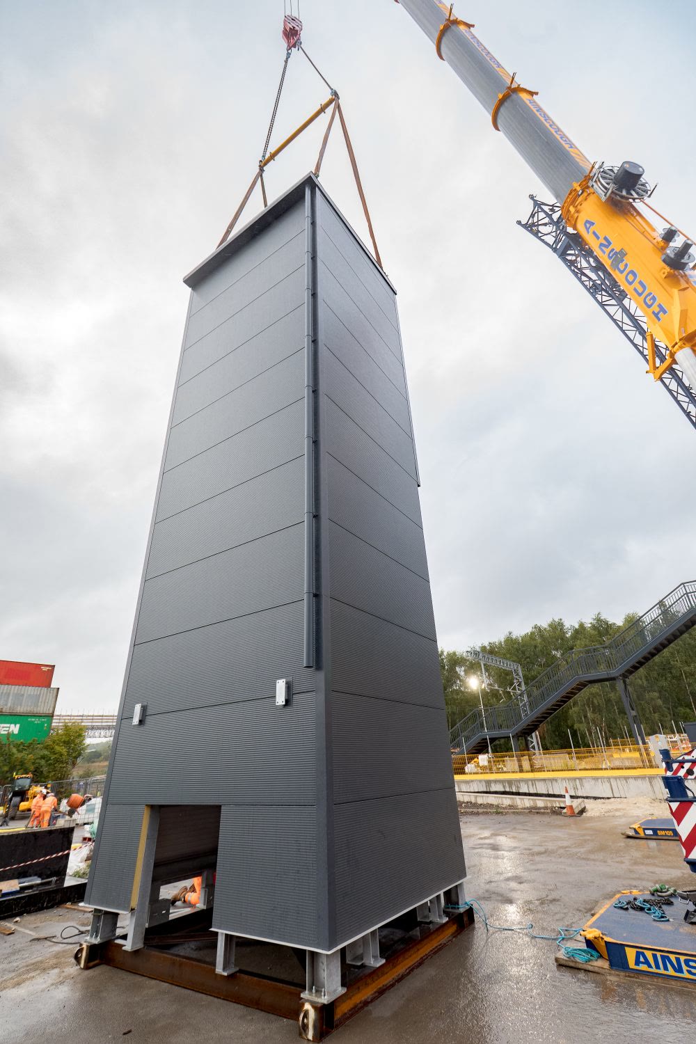 Crane lifting a tall prefabricated lift shaft into position in daylight